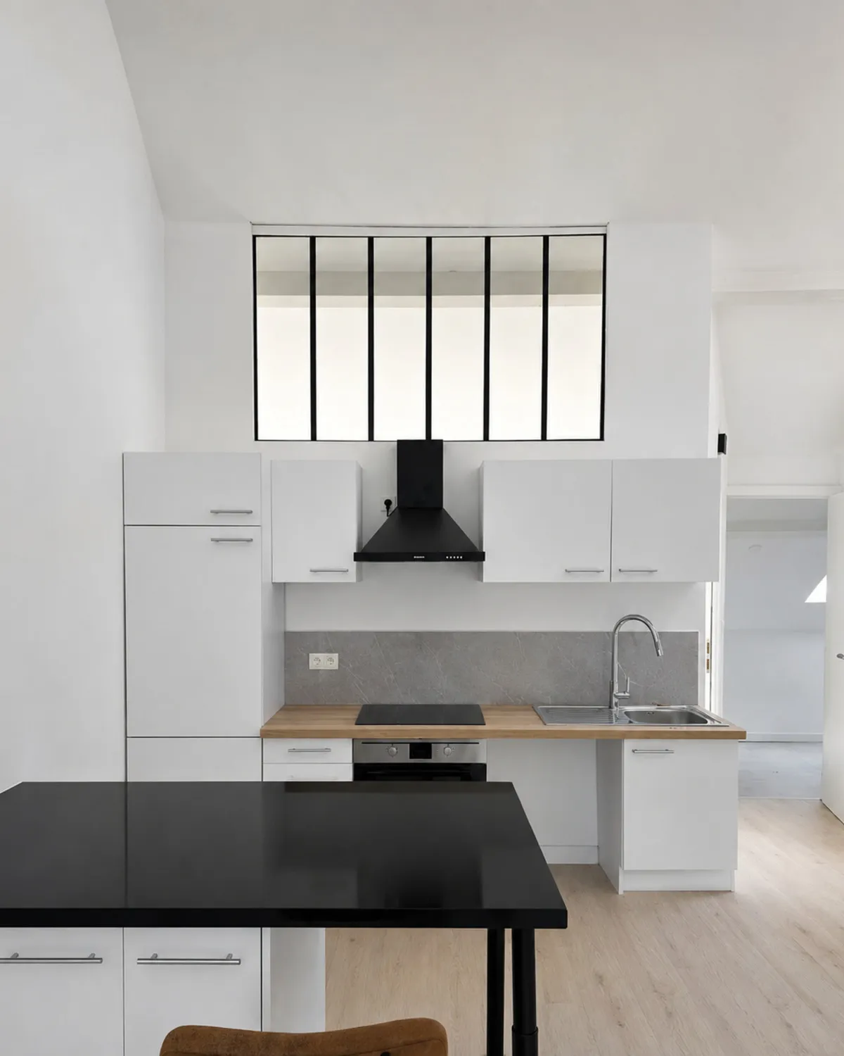 Cuisine aménagée sous comble : meubles blancs, plan de travail en bois, verrière d'atelier et îlot noir.