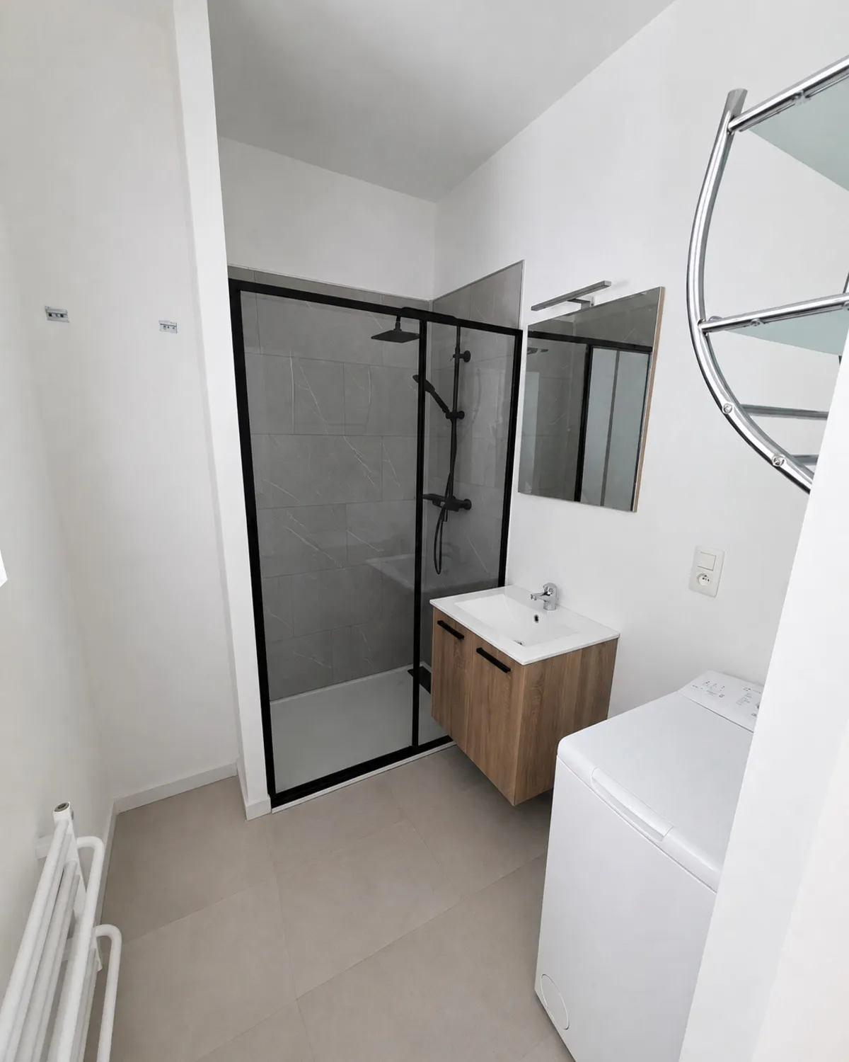 La même salle de bain terminée : douche vitrée à profilés noirs, meuble vasque suspendu et carrelage au sol.